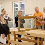 Attēlā redzama piecu cilvēku grupa, kas stāv ap koka darbagaldu. Viens cilvēks tur loga rāmi, kamēr pārējie vēro un pieraksta. Uz darbagalda ir instrumenti un materiāli, tostarp lente un piezīmju grāmatiņas. Vide šķiet kā darbnīca vai klase ar koka grīdām un sienām, logiem fonā un dažiem papildu logu rāmjiem, kas atspiedušies pret sienu.