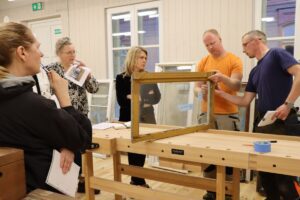 Attēlā redzama piecu cilvēku grupa, kas stāv ap koka darbagaldu. Viens cilvēks tur loga rāmi, kamēr pārējie vēro un pieraksta. Uz darbagalda ir instrumenti un materiāli, tostarp lente un piezīmju grāmatiņas. Vide šķiet kā darbnīca vai klase ar koka grīdām un sienām, logiem fonā un dažiem papildu logu rāmjiem, kas atspiedušies pret sienu.