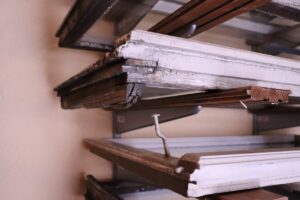 Attēlā redzama kaudze vecu logu rāmju, kas novietoti uz metāla plauktiem. Logu rāmji ir nolietoti, ar nolupušu krāsu un redzamiem plaisām koka materiālā. Dažiem rāmjiem ir stikla paneļu atliekas, kamēr citi ir tukši.