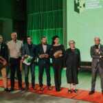 Astoņi cilvēki stāv uz sarkanā paklāja zaļā apgaismojuma fonā; daži tur plāksnes vai sertifikātus, kreisajā pusē redzams 'ROCKWOOL' baneris, labajā pusē ekrāns ar tekstu 'Architecture Construction and Design in Baltics 2025' un āra būves attēlu