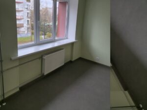 Neliela telpa ar gaiši zaļām sienām un tumšu grīdu, liels balts logs ar palodzi, zem loga balts radiators un metāla caurules gar sienu; ārpus loga redzama daudzdzīvokļu māja un koki