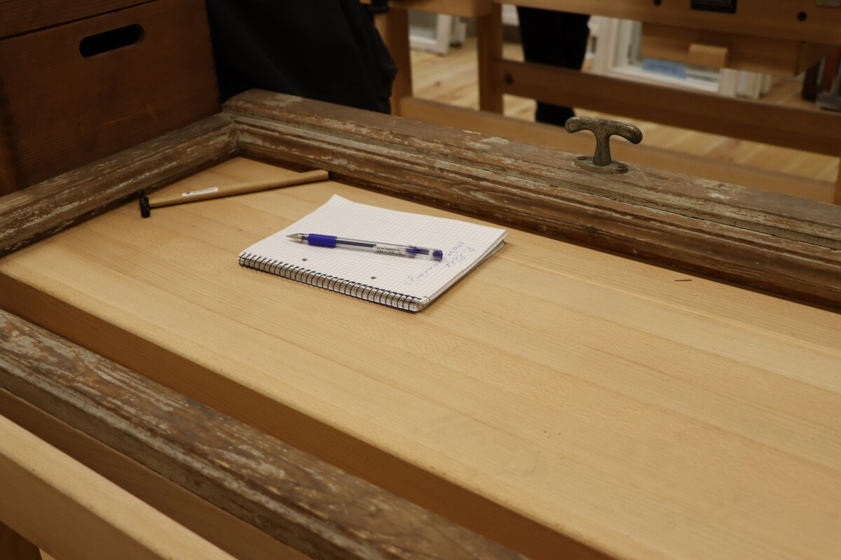 Attēlā redzams koka galds ar vecu, rustisku rāmi ap to. Uz galda ir spirālveida piezīmju grāmatiņa ar grafpapīru un zilu pildspalvu, kas novietota uz tās.