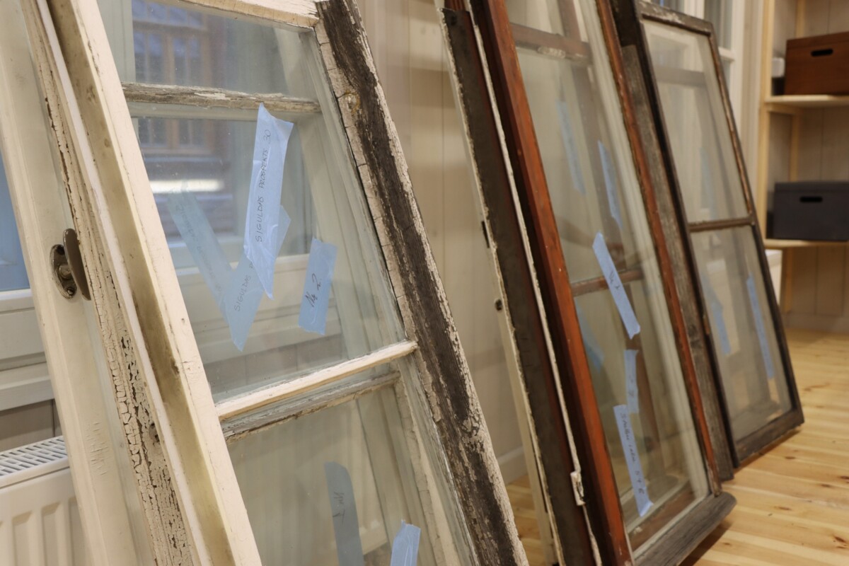 Attēlā redzami vairāki veci logu rāmji, kas atspiedušies pret sienu. Logu rāmji ir dažādās nolietojuma stadijās - daži ar nolupušu krāsu, citi izskatās labāk saglabājušies. Katram loga rāmim ir pievienotas zilas līmlapiņas uz stikla paneļiem.