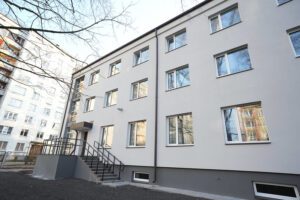 Pašvaldība turpinās uzlabošanas darbus Rīgas Hokeja skolas ēkās -Moderna trīsstāvu daudzdzīvokļu ēka ar gaišu fasādi un vairākām logu rindām; pie ieejas kāpnes, apkārtnē redzama vēl viena līdzīga ēka un kaili koki