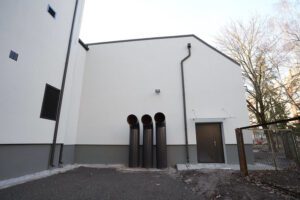 Pašvaldība turpinās uzlabošanas darbus Rīgas Hokeja skolas ēkās - Ēkas fasāde ar gaišu apdari un trim vertikāliem melniem cauruļvadiem pie sienas; pa labi – melnas durvis ar apgaismes ķermeni virs tām, priekšplānā asfalts, fonā koki un cita ēka