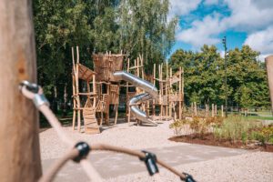 Koka rotaļu laukuma konstrukcija ar vairākiem līmeņiem, kāpšanas platformām un metāla spirāles slidkalniņu centrā. Apkārt redzami koki, grants segums un dabiskas koka kāpelēšanas struktūras, bet priekšplānā — virvju elements, kas iezīmē laukuma tuvplānu. Skaidra, saulaina diena ar gaišām debesīm un mākoņiem.