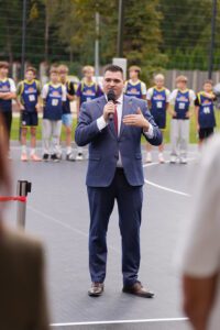 Persona uzvalkā runā mikrofonā āra sporta laukumā; fonā redzami jaunieši sporta formās