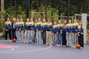 Cilvēku grupa stāv rindā uz āra basketbola laukuma, tērpti vienādās zilās un dzeltenās sporta formās ar logotipiem; abos rindas galos uz zemes novietotas basketbola bumbas, fonā redzams žogs un zaļumi