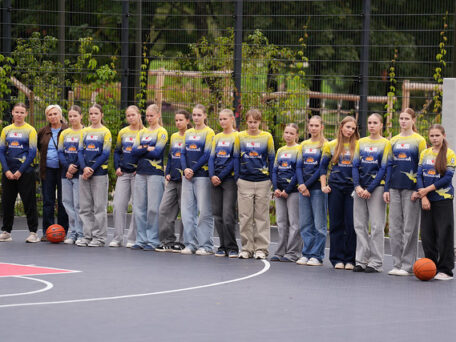 Cilvēku grupa stāv rindā uz āra basketbola laukuma, tērpti vienādās zilās un dzeltenās sporta formās ar logotipiem; abos rindas galos uz zemes novietotas basketbola bumbas, fonā redzams žogs un zaļumi