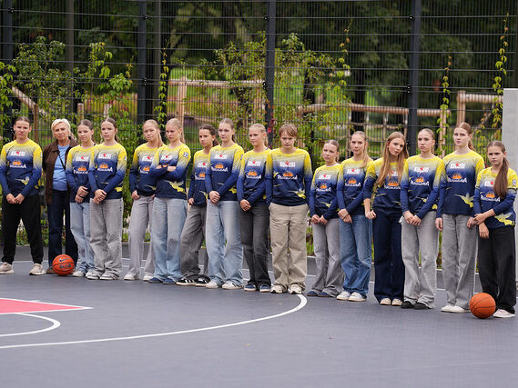 Cilvēku grupa stāv rindā uz āra basketbola laukuma, tērpti vienādās zilās un dzeltenās sporta formās ar logotipiem; abos rindas galos uz zemes novietotas basketbola bumbas, fonā redzams žogs un zaļumi