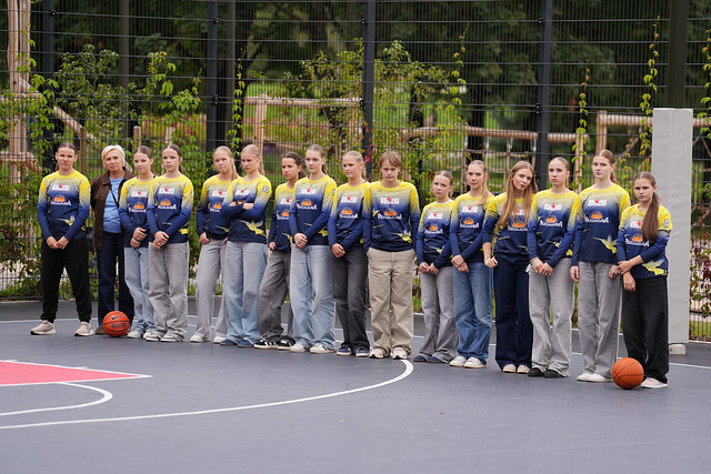Cilvēku grupa stāv rindā uz āra basketbola laukuma, tērpti vienādās zilās un dzeltenās sporta formās ar logotipiem; abos rindas galos uz zemes novietotas basketbola bumbas, fonā redzams žogs un zaļumi