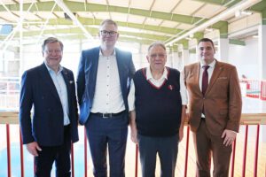 Grupa ar četriem cilvēkiem stāv iekštelpās uz paaugstinājuma ar sarkanu margu priekšplānā. Visi ir tērpušies formālā vai pusformālā apģērbā – uzvalkos vai vestē ar kreklu. Fonā redzama plaša sporta vai industriāla tipa zāle ar augstiem griestiem, metāla sijām un lieliem logiem, kas ielaiž daudz dabiskās gaismas.