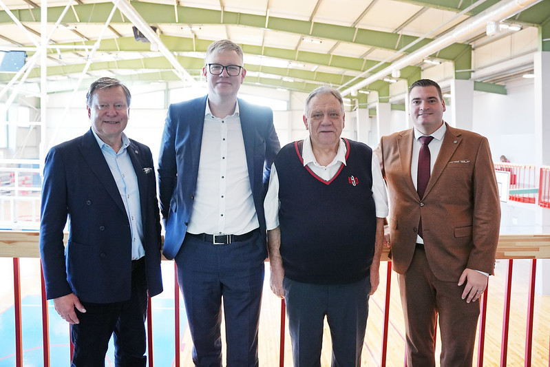 Grupa ar četriem cilvēkiem stāv iekštelpās uz paaugstinājuma ar sarkanu margu priekšplānā. Visi ir tērpušies formālā vai pusformālā apģērbā – uzvalkos vai vestē ar kreklu. Fonā redzama plaša sporta vai industriāla tipa zāle ar augstiem griestiem, metāla sijām un lieliem logiem, kas ielaiž daudz dabiskās gaismas.