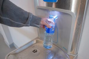 Rokturā turēta plastmasas pudele tiek piepildīta ar ūdeni pie sienā iebūvēta ūdens uzpildes aparāta. No augšējās sprauslas ieplūst ūdens, ko izgaismo zila LED gaisma. Pudele ir novietota uz metāla virsmas ar notekcaurumu, un fonā redzama gaiša siena un grīda.