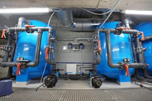 Peldbaseina tehniskā telpa ar diviem lieliem zilas krāsas ūdens filtrācijas rezervuāriem, savienotiem ar cauruļvadiem, vārstiem un sūkņiem; redzama ventilācijas sistēma un industriāls grīdas režģis