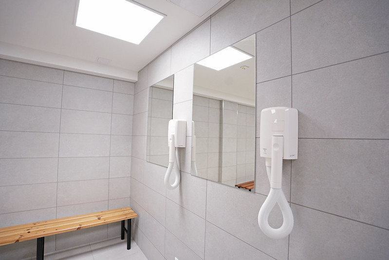 Pārģērbšanās telpa ar gaiši pelēkām flīzēm, sienā iebūvētu spoguli, pie sienas piestiprinātu matu fēnu un koka soliņu