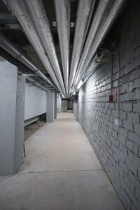 Tehniskais koridors peldbaseina ēkā ar griestiem izvietotām siltinātām caurulēm, betona grīdu un ķieģeļu sienu; redzama inženierkomunikāciju infrastruktūra