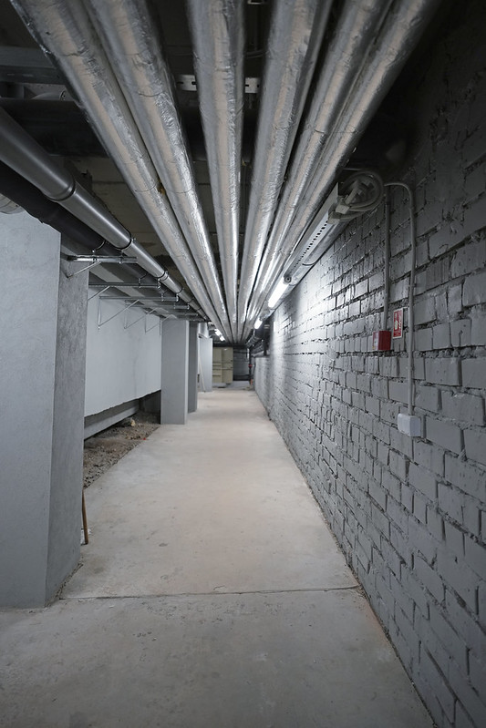 Tehniskais koridors peldbaseina ēkā ar griestiem izvietotām siltinātām caurulēm, betona grīdu un ķieģeļu sienu; redzama inženierkomunikāciju infrastruktūra