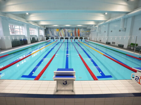 Iekštelpu peldbaseins ar vairākām peldēšanas celiņu rindām, starta blokiem baseina malā un karodziņiem virs ūdens; gaiša, renovēta telpa ar augstiem griestiem.