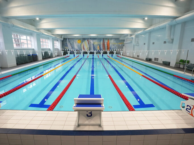 Iekštelpu peldbaseins ar vairākām peldēšanas celiņu rindām, starta blokiem baseina malā un karodziņiem virs ūdens; gaiša, renovēta telpa ar augstiem griestiem.