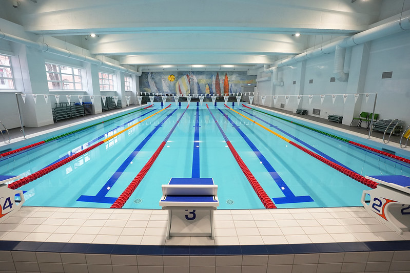 Iekštelpu peldbaseins ar vairākām peldēšanas celiņu rindām, starta blokiem baseina malā un karodziņiem virs ūdens; gaiša, renovēta telpa ar augstiem griestiem.