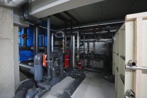Peldbaseina tehniskā telpa ar cauruļvadu sistēmu, sūkņiem un siltummezgla iekārtām; redzamas metāla un plastmasas caurules, vārsti un inženiertehniskais aprīkojums