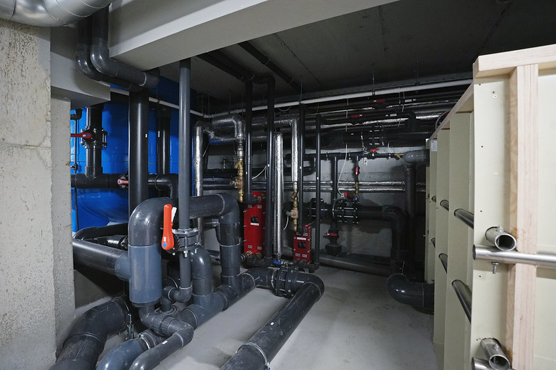 Peldbaseina tehniskā telpa ar cauruļvadu sistēmu, sūkņiem un siltummezgla iekārtām; redzamas metāla un plastmasas caurules, vārsti un inženiertehniskais aprīkojums