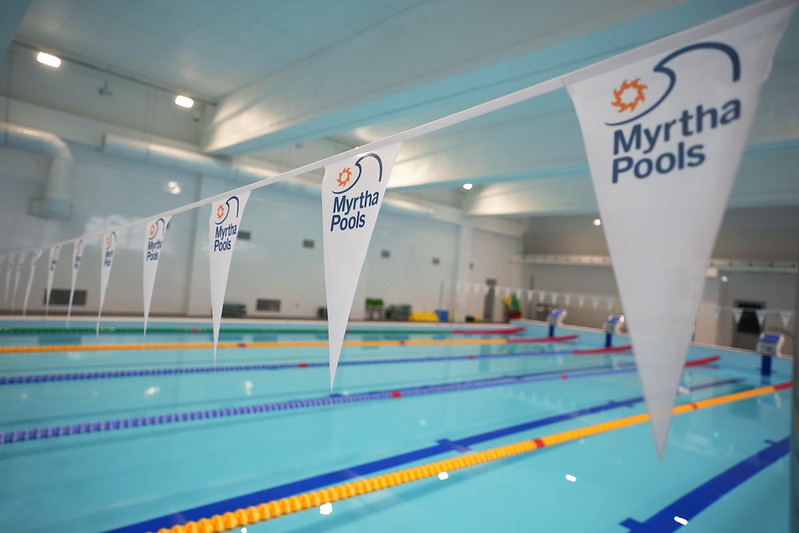 Iekštelpu peldbaseins ar vairākām peldēšanas celiņu rindām, starta blokiem baseina malā un karodziņiem virs ūdens; gaiša, renovēta telpa ar augstiem griestiem