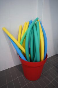 Sarkanā traukā novietotas peldēšanas nūdeles dažādās krāsās (zilā, zaļā un dzeltenā), kas paredzētas ūdens nodarbībām baseinā; inventārs novietots pie flīzētas sienas