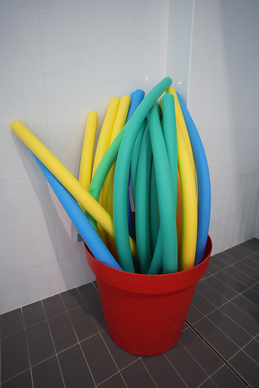 Sarkanā traukā novietotas peldēšanas nūdeles dažādās krāsās (zilā, zaļā un dzeltenā), kas paredzētas ūdens nodarbībām baseinā; inventārs novietots pie flīzētas sienas