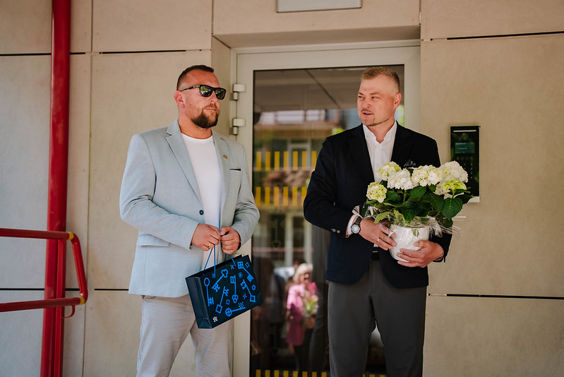 Divi cilvēki stāv pie ēkas stikla durvīm; viens tur rokās baltu ziedu pušķi, otram rokās tumša dāvanu maisiņš; fonā redzama gaiša fasāde un sarkanas margas