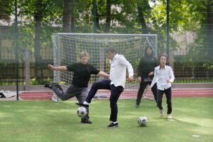 Skolēni spēlē futbolu āra sporta laukumā, redzami vārti un vairāki spēlētāji cīņā par bumbu.