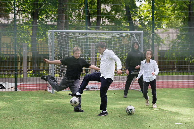 Skolēni spēlē futbolu āra sporta laukumā, redzami vārti un vairāki spēlētāji cīņā par bumbu.
