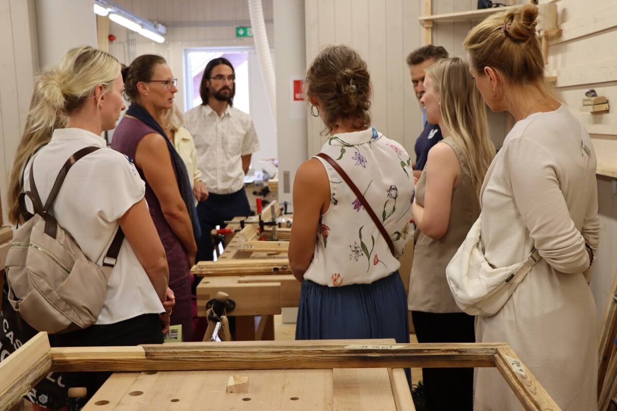 Grupa cilvēku darbnīcas vai mācību telpā pulcējas ap koka darba galdiem; uz galdiem redzami dažādi instrumenti un aprīkojums, telpa ir gaiša ar baltām sienām un dienasgaismas apgaismojumu