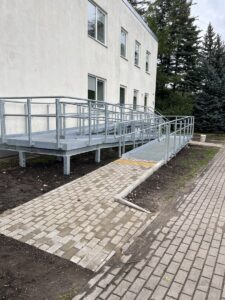 Pieejama tualetes telpa ar zaļām flīzēm, tualeti, atbalsta rokturiem, roku žāvētāju un papīra turētāju