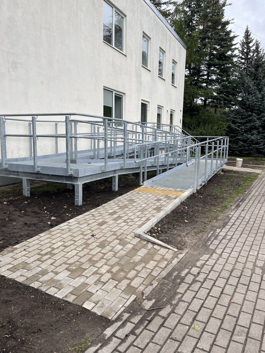 Pieejama tualetes telpa ar zaļām flīzēm, tualeti, atbalsta rokturiem, roku žāvētāju un papīra turētāju