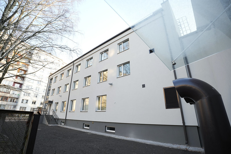 Pašvaldība turpinās uzlabošanas darbus Rīgas Hokeja skolas ēkās - Trīsstāvu ēka ar gaišu fasādi un vairākām logu rindām; priekšplānā metāla žogs un bruģēta teritorija, pa kreisi koks un kāpnes uz ieeju, fonā redzama cita daudzstāvu ēka.