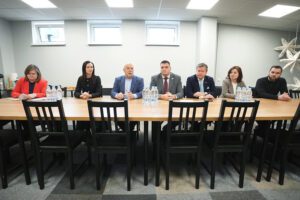 Pašvaldība turpinās uzlabošanas darbus Rīgas Hokeja skolas ēkās - Sanāksmes telpa ar septiņiem cilvēkiem pie gara koka galda; fonā trīs logi, uz galda ūdens pudeles, telpa ar mūsdienīgu apgaismojumu un dekoratīviem elementiem