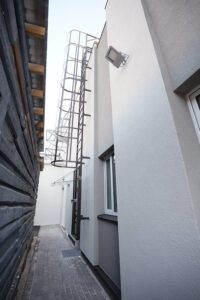 Pašvaldība turpinās uzlabošanas darbus Rīgas Hokeja skolas ēkās - Šaura ejas telpa starp divām ēkām – labajā pusē moderna ēka ar pelēku fasādi, logu un metāla kāpnēm uz jumta; kreisajā pusē koka apšuvuma siena, bruģēta zeme un āra apgaismojums