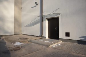 Pašvaldība turpinās uzlabošanas darbus Rīgas Hokeja skolas ēkās - Ēkas fasāde ar brūnām durvīm un stikla jumtiņu virs tām; pie sienas redzamas divas novērošanas kameras un āra apgaismojums, priekšā neliels uzbrauktuves panduss un sniega paliekas.