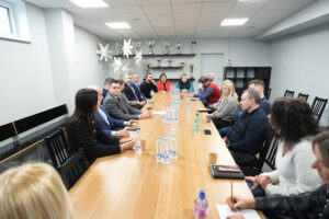 Pašvaldība turpinās uzlabošanas darbus Rīgas Hokeja skolas ēkās - Sanāksmes telpa ar apmēram 15 cilvēkiem pie gara koka galda; uz galda ūdens pudeles un krūzes, fonā plaukti ar trofejām un zvaigžņu formas dekori pie griestiem