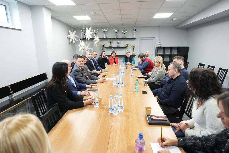Pašvaldība turpinās uzlabošanas darbus Rīgas Hokeja skolas ēkās - Sanāksmes telpa ar apmēram 15 cilvēkiem pie gara koka galda; uz galda ūdens pudeles un krūzes, fonā plaukti ar trofejām un zvaigžņu formas dekori pie griestiem