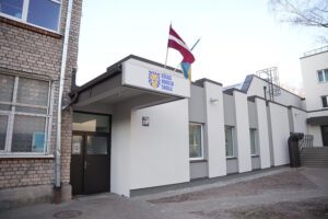 Pašvaldība turpinās uzlabošanas darbus Rīgas Hokeja skolas ēkās - Ēkas fasāde ar uzrakstu 'Rīgas Hokeja Skola', Latvijas un zilu-dzeltenu karogu uz jumta, baltiem mūsdienīgiem sienu paneļiem, lieliem logiem un pievienotu vecāku ķieģeļu daļu. Pie ieejas redzams numurs '159'.