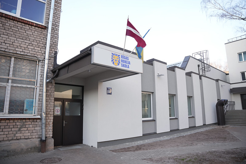 Pašvaldība turpinās uzlabošanas darbus Rīgas Hokeja skolas ēkās - Ēkas fasāde ar uzrakstu 'Rīgas Hokeja Skola', Latvijas un zilu-dzeltenu karogu uz jumta, baltiem mūsdienīgiem sienu paneļiem, lieliem logiem un pievienotu vecāku ķieģeļu daļu. Pie ieejas redzams numurs '159'.