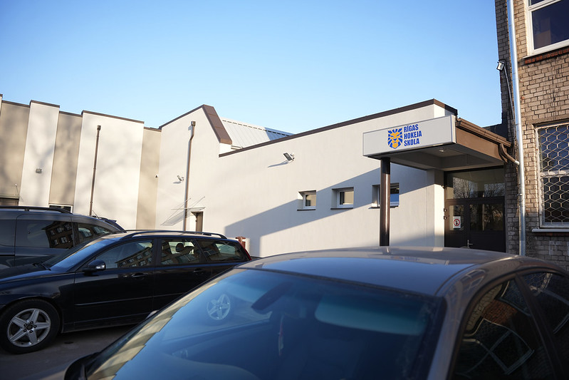 Pašvaldība turpinās uzlabošanas darbus Rīgas Hokeja skolas ēkās - Autostāvvieta ar vairākām automašīnām pie modernas ēkas ar uzrakstu 'Rīgas Hokeja Skola' zem skaidrām zilām debesīm