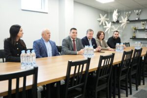 Pašvaldība turpinās uzlabošanas darbus Rīgas Hokeja skolas ēkās - Piecu cilvēku sanāksme pie gara koka galda; uz galda ūdens pudeles, fonā plaukti ar trofejām un zvaigžņu formas dekori pie griestiem
