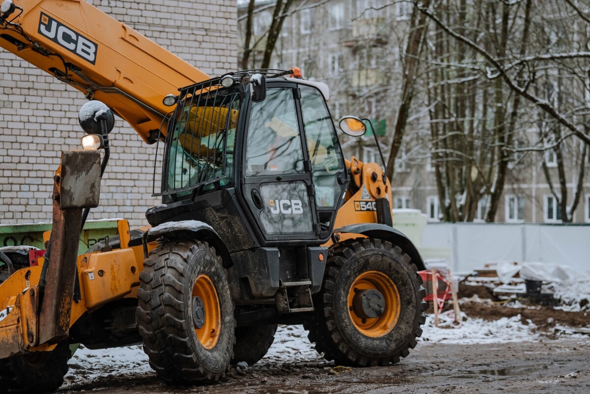 Dzelteni melns JCB 540-140 teleskopiskais iekrāvējs būvlaukumā, fonā ķieģeļu ēka, koki ar sniegu un dažādi būvmateriāli