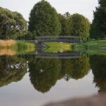 Mazs tiltiņš pār mierīgu ūdenstilpni parkā, kurā atspoguļojas apkārtējie koki un tilts; apkārtne ir zaļa un klusa.