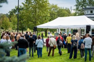 Cilvēku grupa pulcējas zem baltas nojumes āra pasākumā parkā; fonā redzama zaļa zāle un koki, uzstādīti skaļruņi norāda uz prezentāciju vai runu