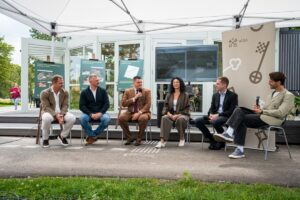 Seši cilvēki sēž uz krēsliem zem baltas nojumes āra pasākumā; fonā redzami stendi ar uzrakstiem 'UZVARAS PARKS' un 'RIGA', apkārt redzama zaļa vide